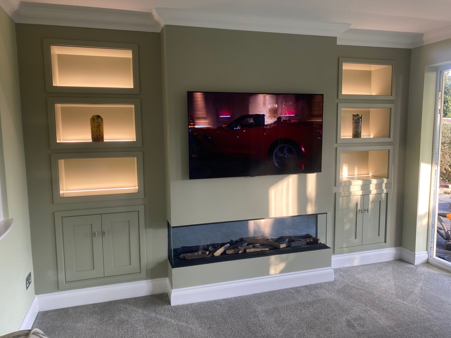 Living room with built-in shelving, TV, and fireplace; sage green walls; illuminated shelves.