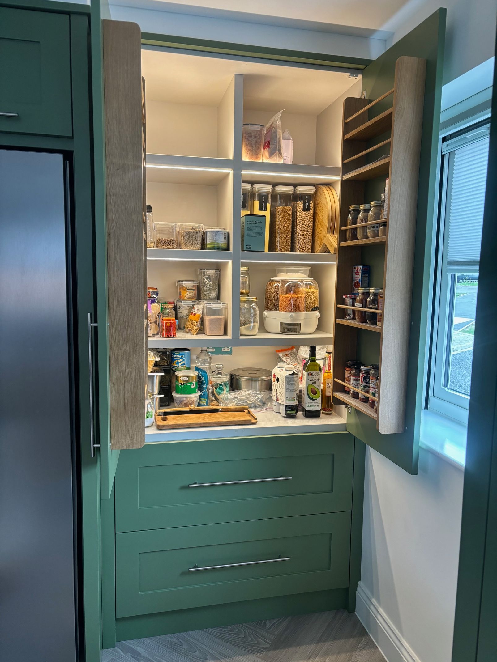 Green pantry cabinet open, showing shelves with food and spices, built-in to kitchen next to refrigerator and window.