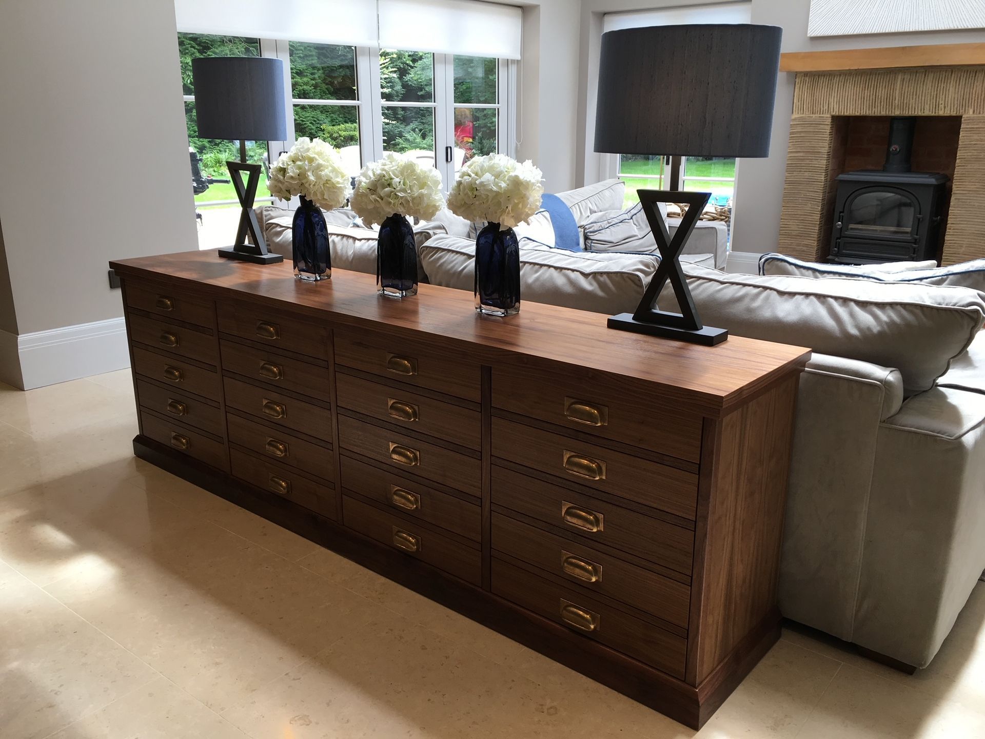 Wooden cabinet with drawers, topped with flowers in blue vases, lamps. Sofa, fireplace in background.