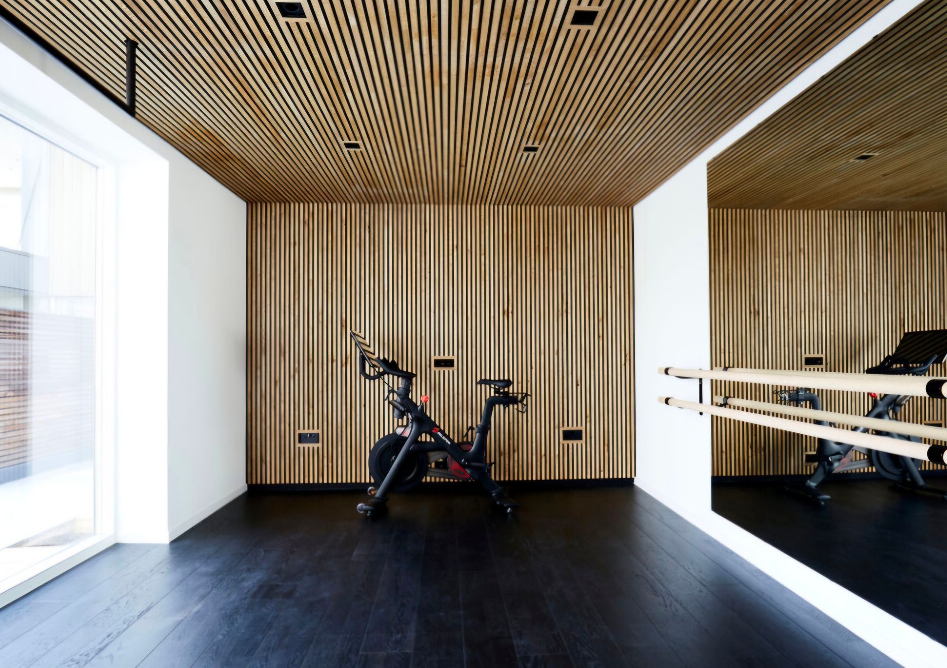Wooden-walled home gym with a spin bike, a mirror with a ballet barre, and a dark wooden floor.