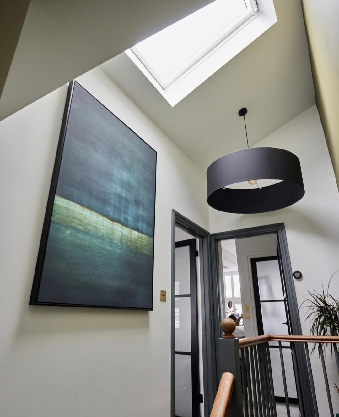 Hallway with large abstract painting, skylight, black pendant light, and dark gray trim.