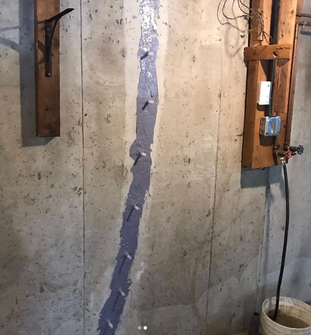 Concrete wall with a repaired vertical crack, filled with gray sealant and nails. Electrical fixtures and a bucket are also visible.
