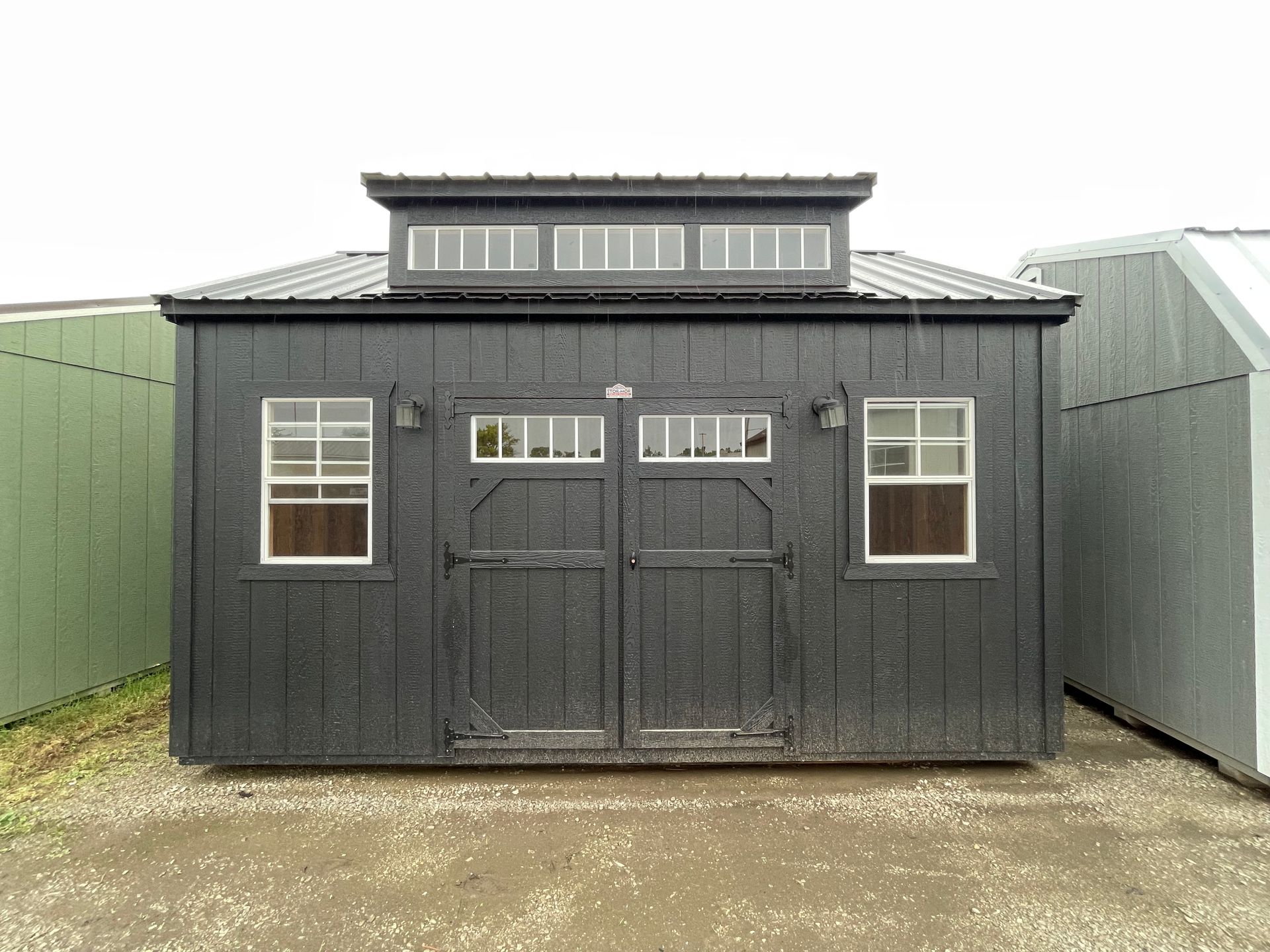 A black shed with a roof and windows is sitting on top of a dirt field.