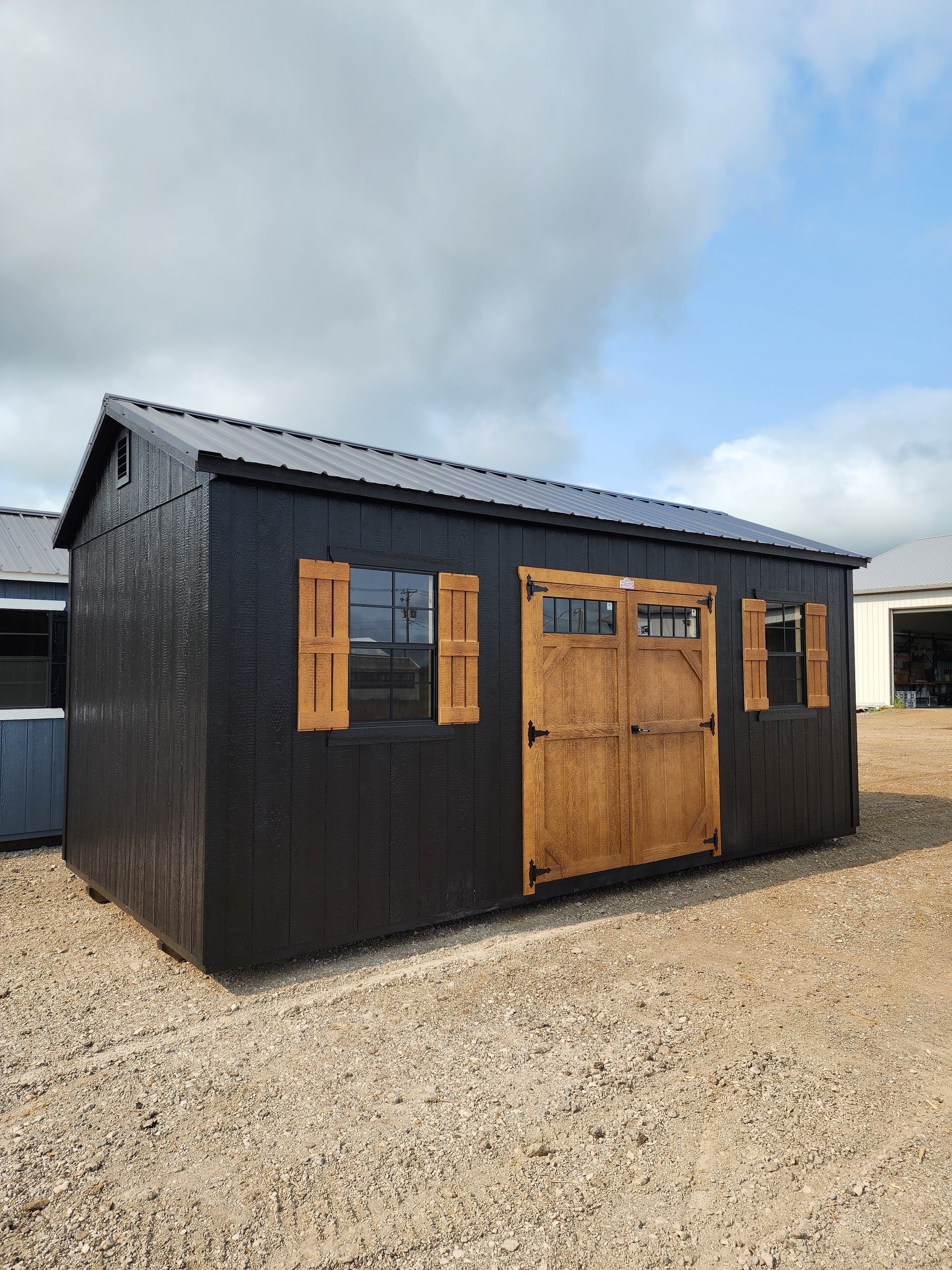 A black shed with wooden doors and windows is sitting on top of a gravel lot.