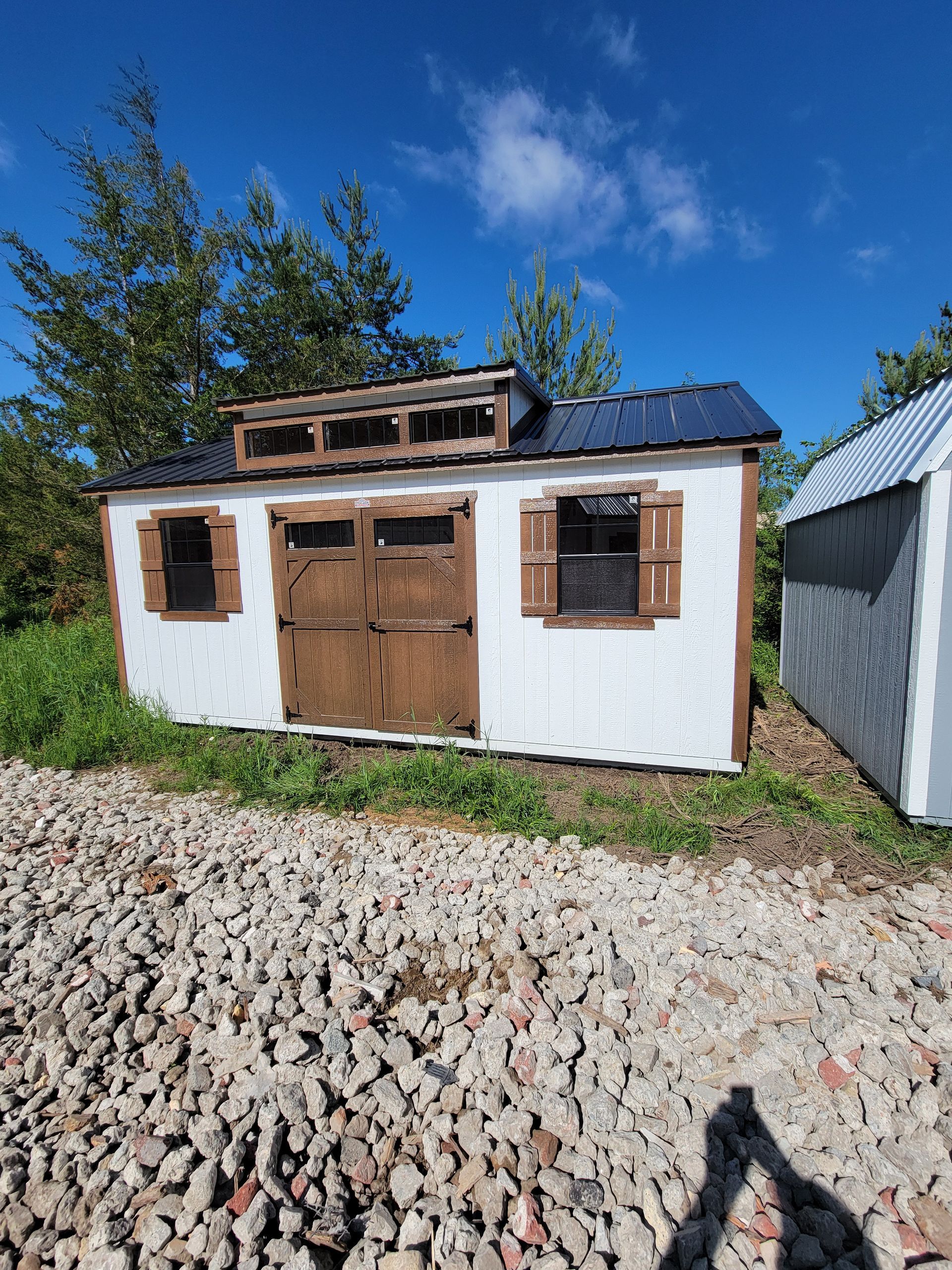 A gray and white shed with shutters on the windows is sitting in the middle of a grassy field.