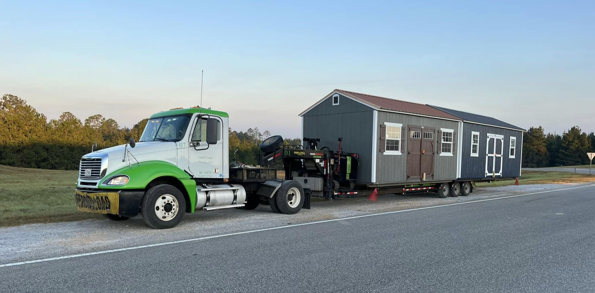 A green and white semi truck is towing a shed down a road.