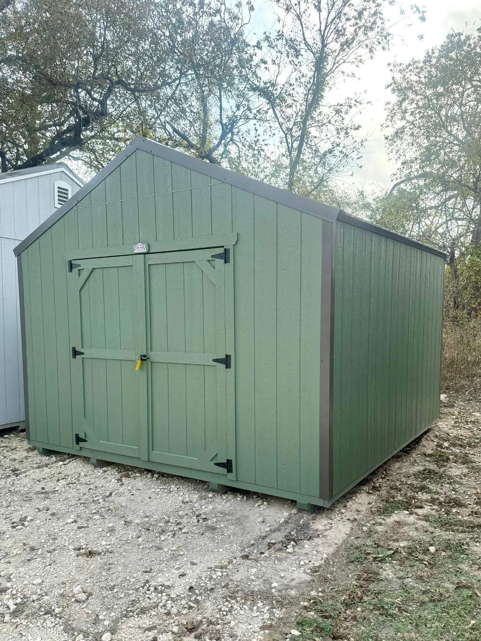 Green wooden shed with double doors on a gravel lot, trees in the background.