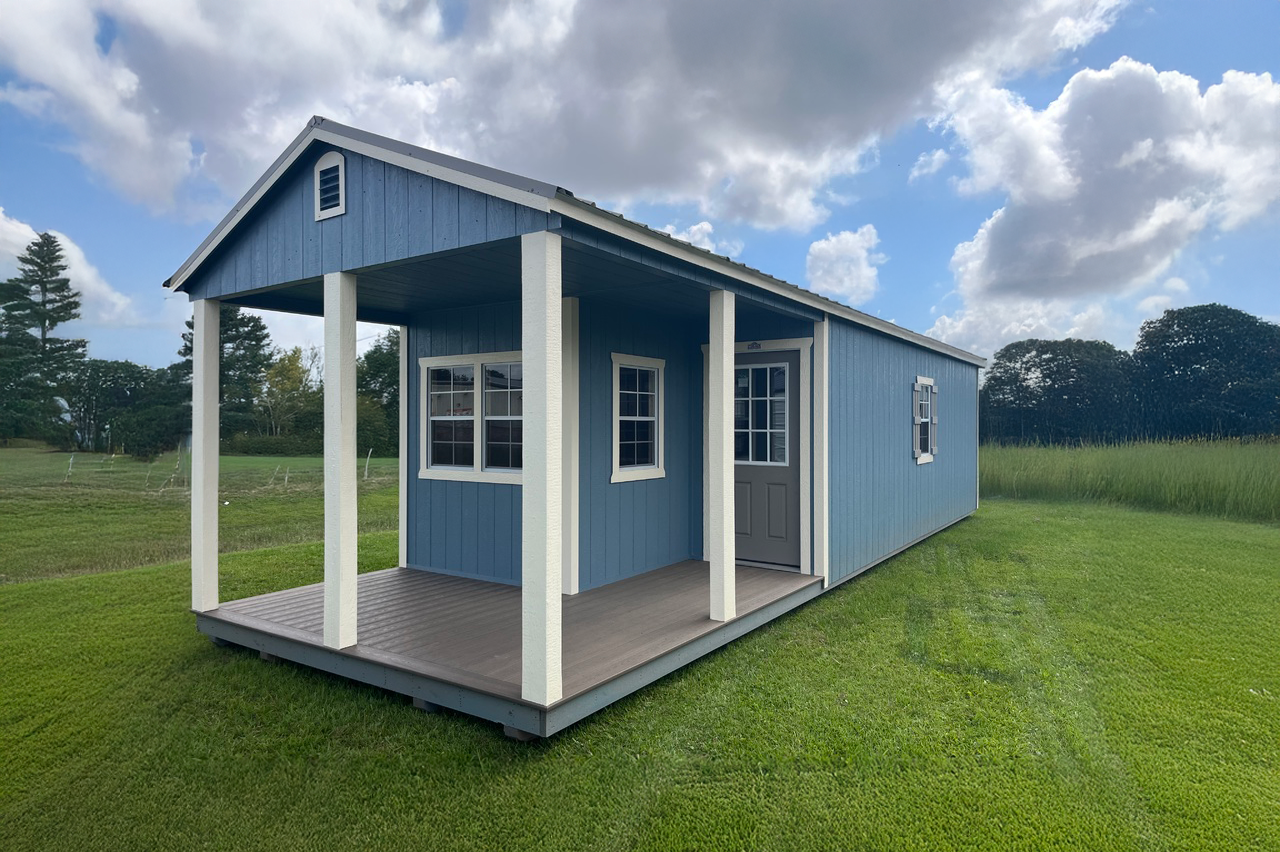 A blue cabin shed with a porch and windows is sitting on top of a lush green field.
