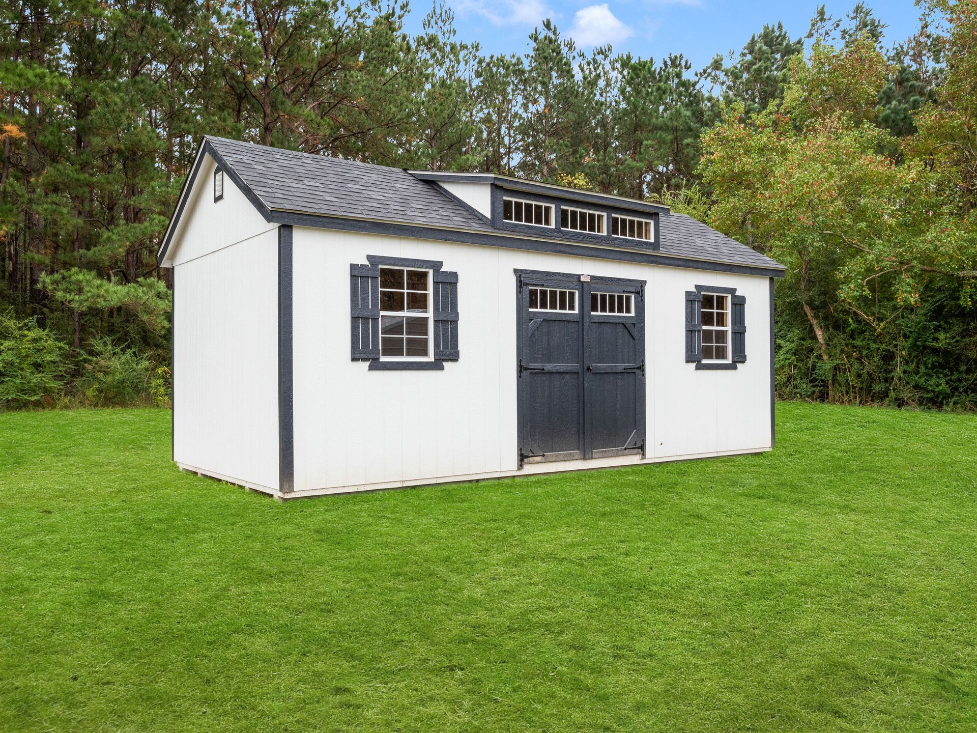 A white shed with black shutters is sitting in the middle of a lush green field.