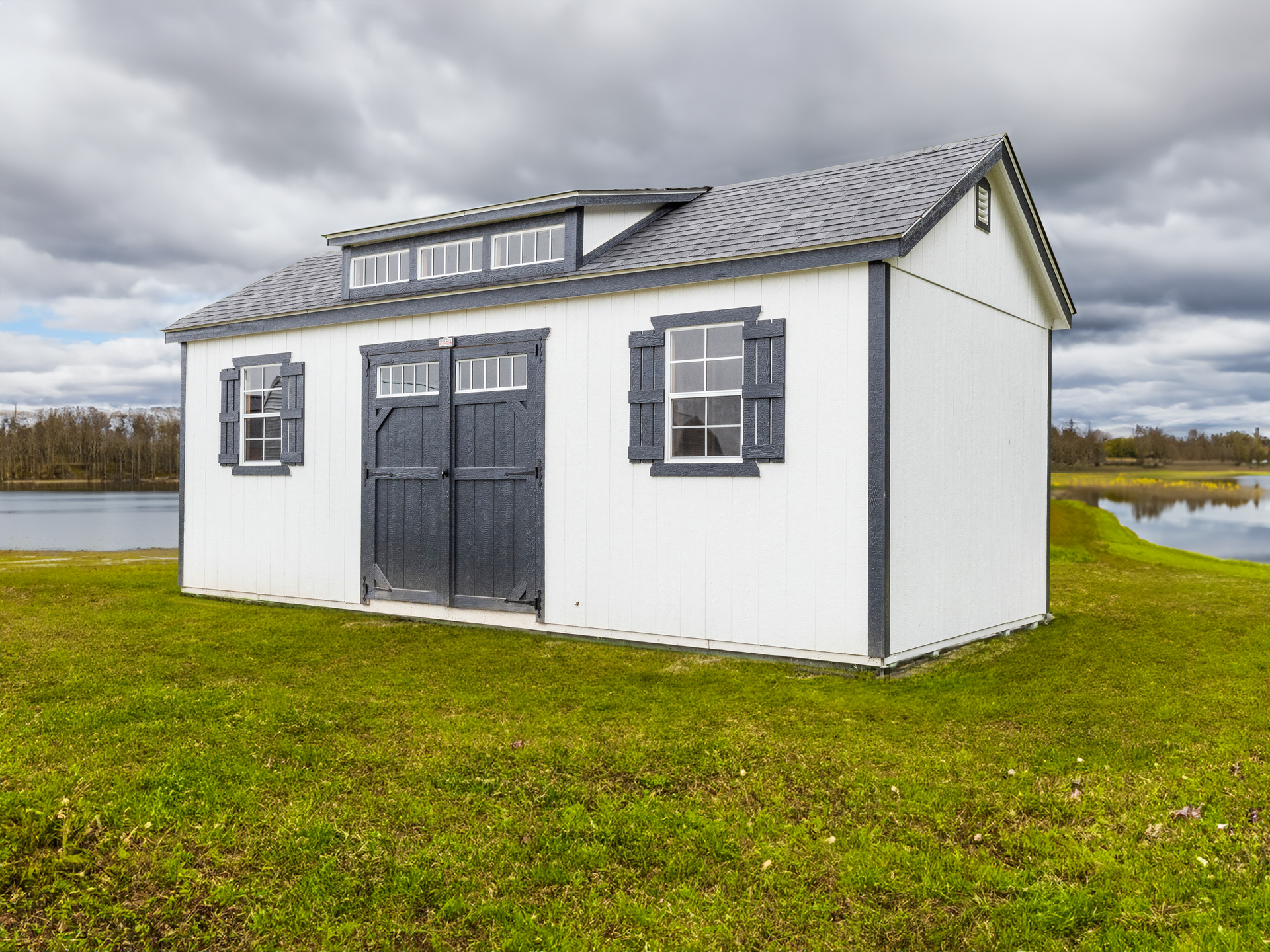 A white shed with black shutters is sitting in the middle of a grassy field next to a body of water.