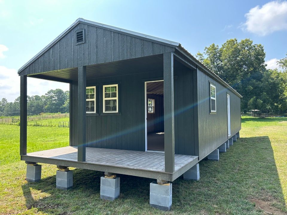 A small black shed with a porch is sitting in the middle of a grassy field.