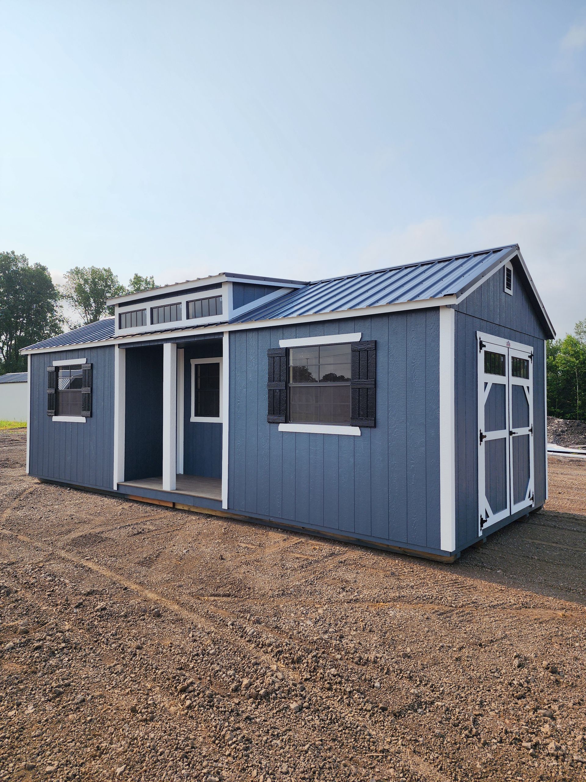 A blue shed with a porch and windows is sitting on top of a pile of gravel.