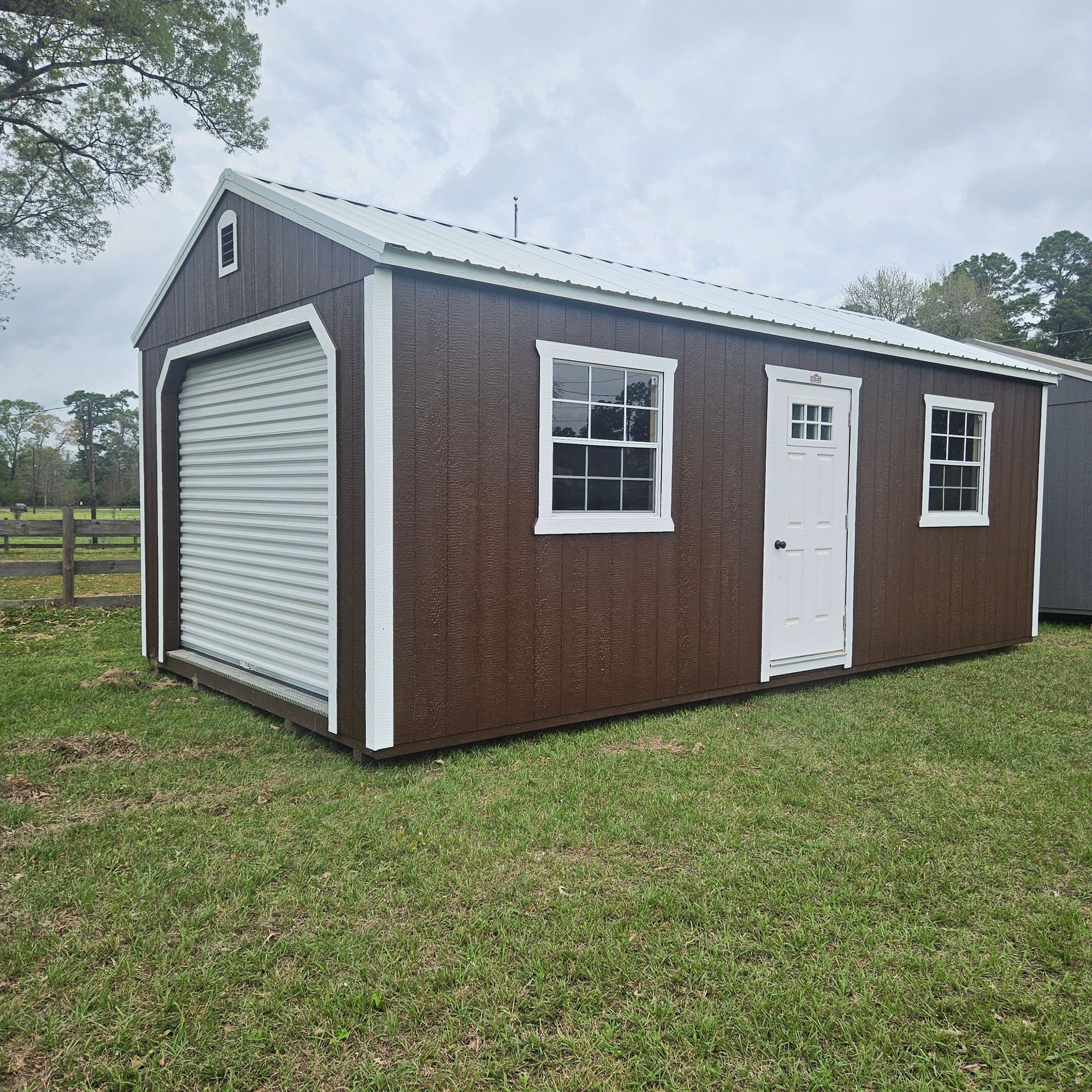 A brown shed with a white door and windows is in the middle of a grassy field.