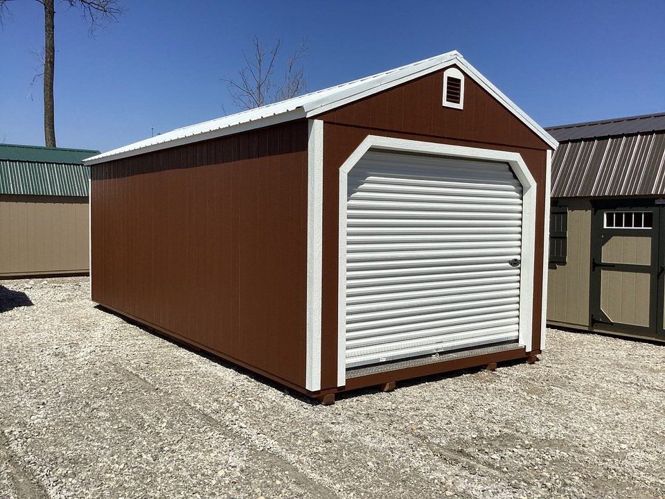 A brown shed with a white garage door is sitting in a gravel lot.