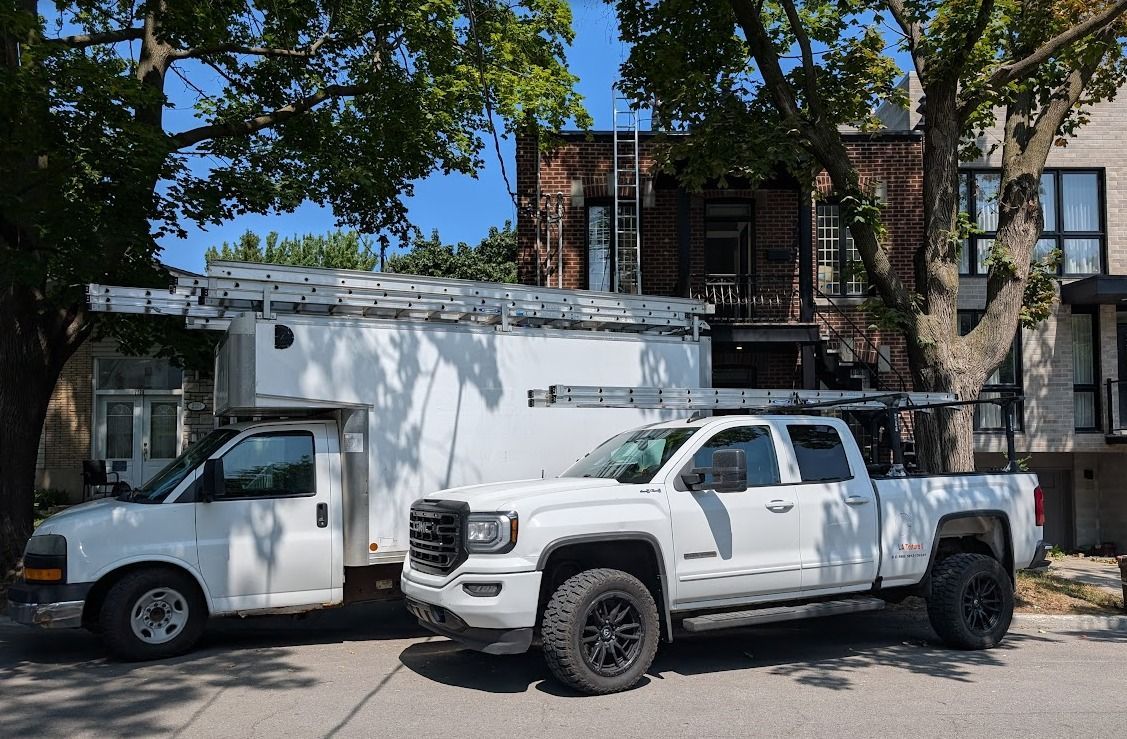 Une camionnette blanche et un pick-up sont stationnés dans une rue, devant un bâtiment en briques, sous des arbres.