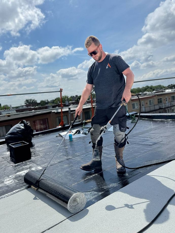 Un ouvrier inspecte un toit plat sous un ciel nuageux, portant un équipement de sécurité et tenant un outil.