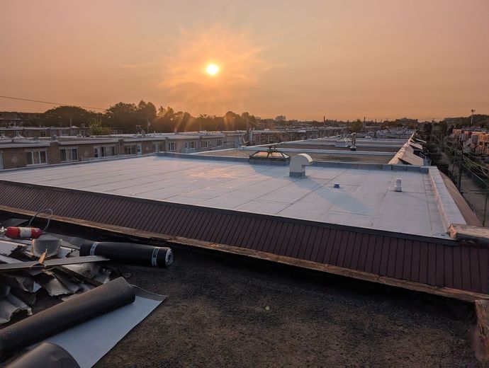 Toiture au coucher du soleil avec panneaux solaires et matériaux de construction, ciel orangé au-dessus des arbres et des bâtiments au loin
