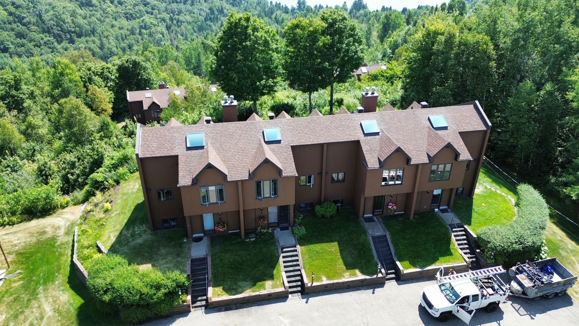Maisons de ville marron avec escaliers et pelouses vertes, entourées d'arbres. Un camion blanc est garé devant.