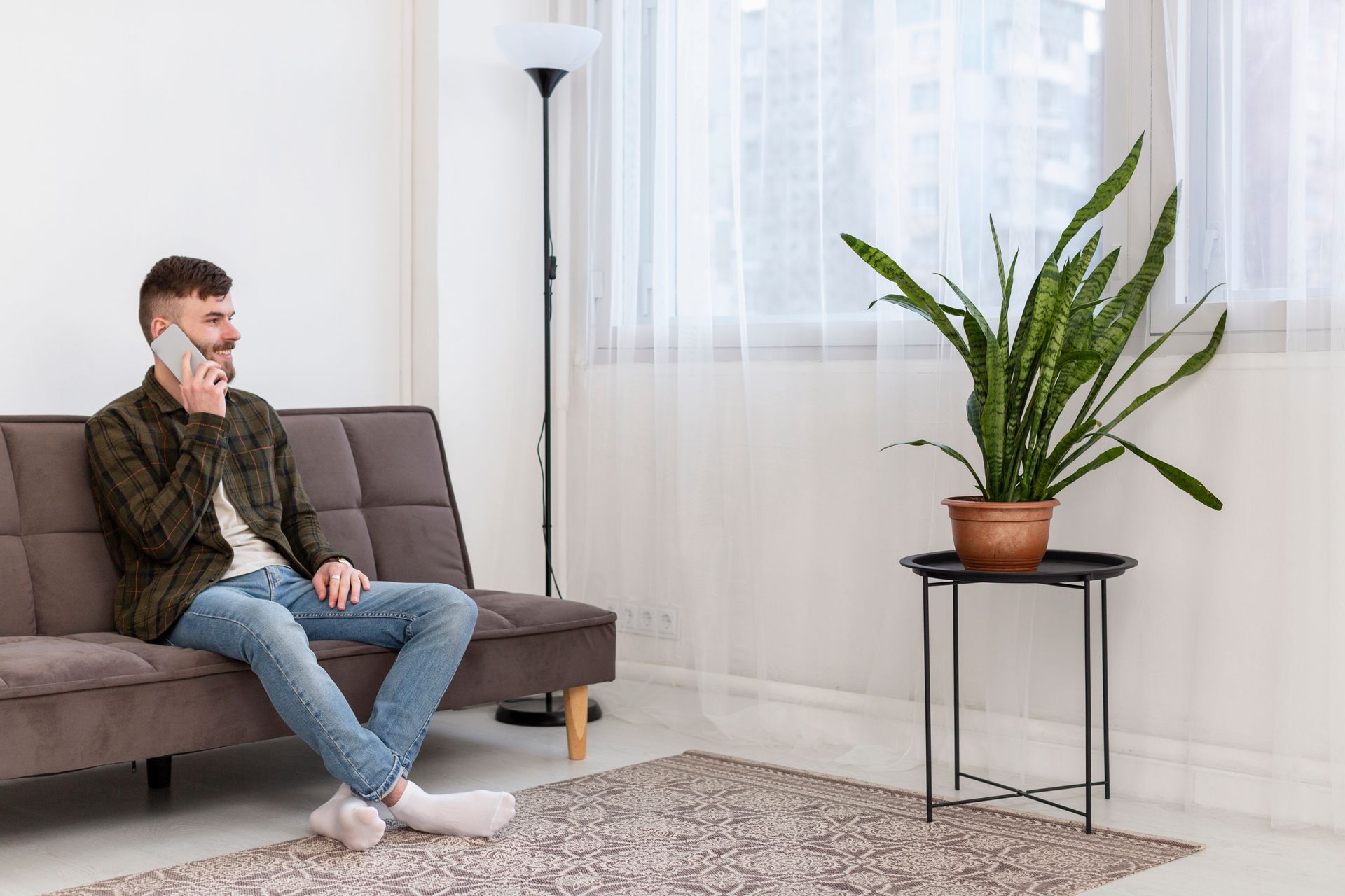 A person wearing a plaid shirt and jeans sits on a brown sofa, talking on a phone, next to a potted plant on a stand.