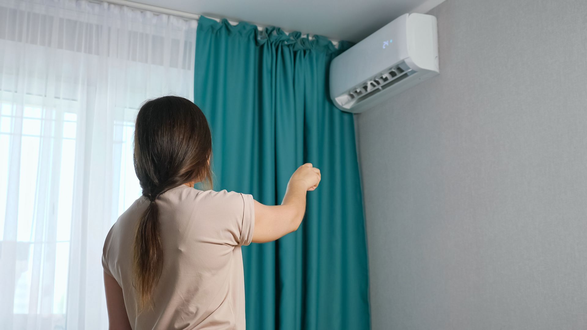 Person pointing a remote at a wall-mounted air conditioner indoors.