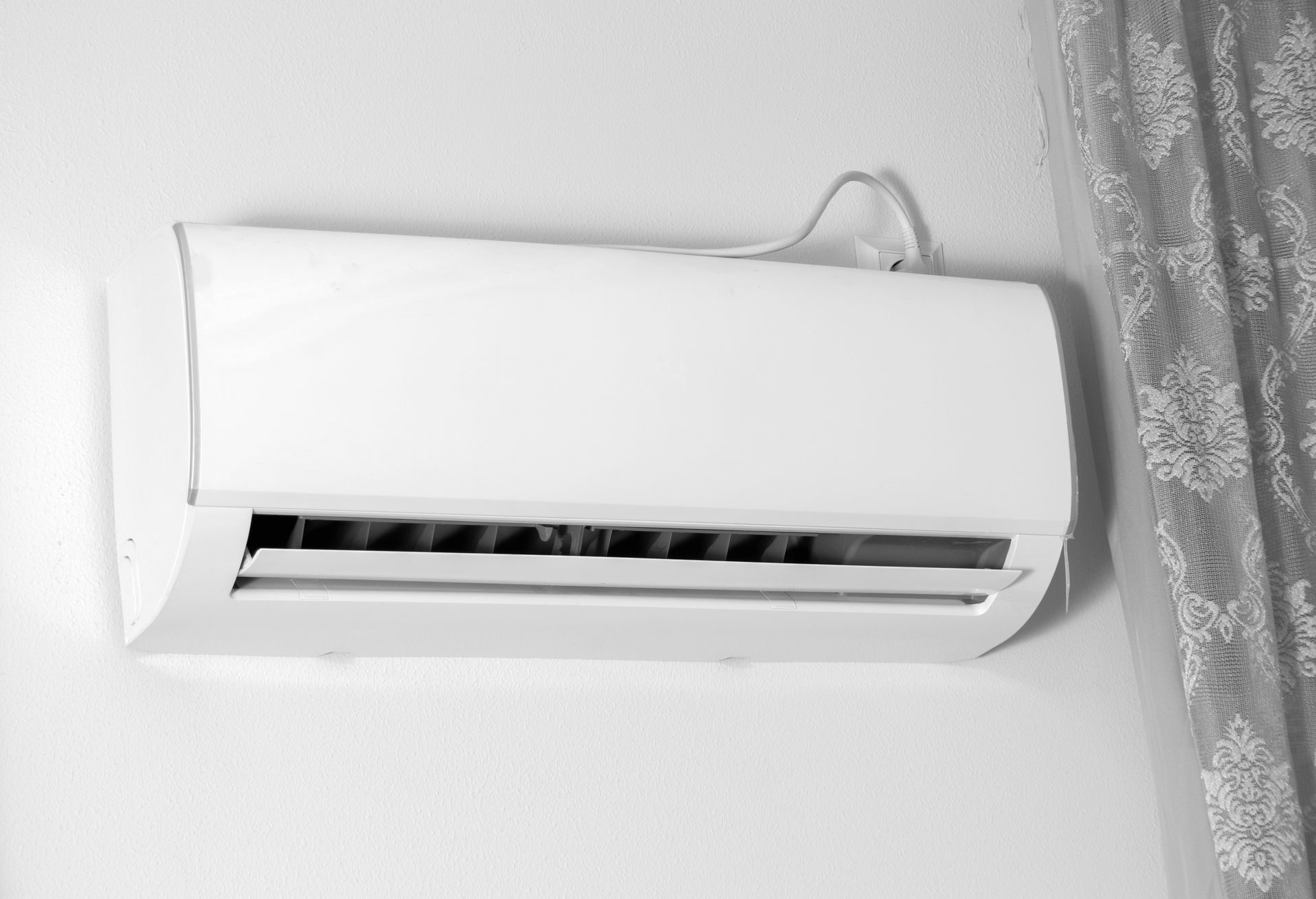 A white wall-mounted air conditioner unit is installed on a white wall next to a patterned curtain.