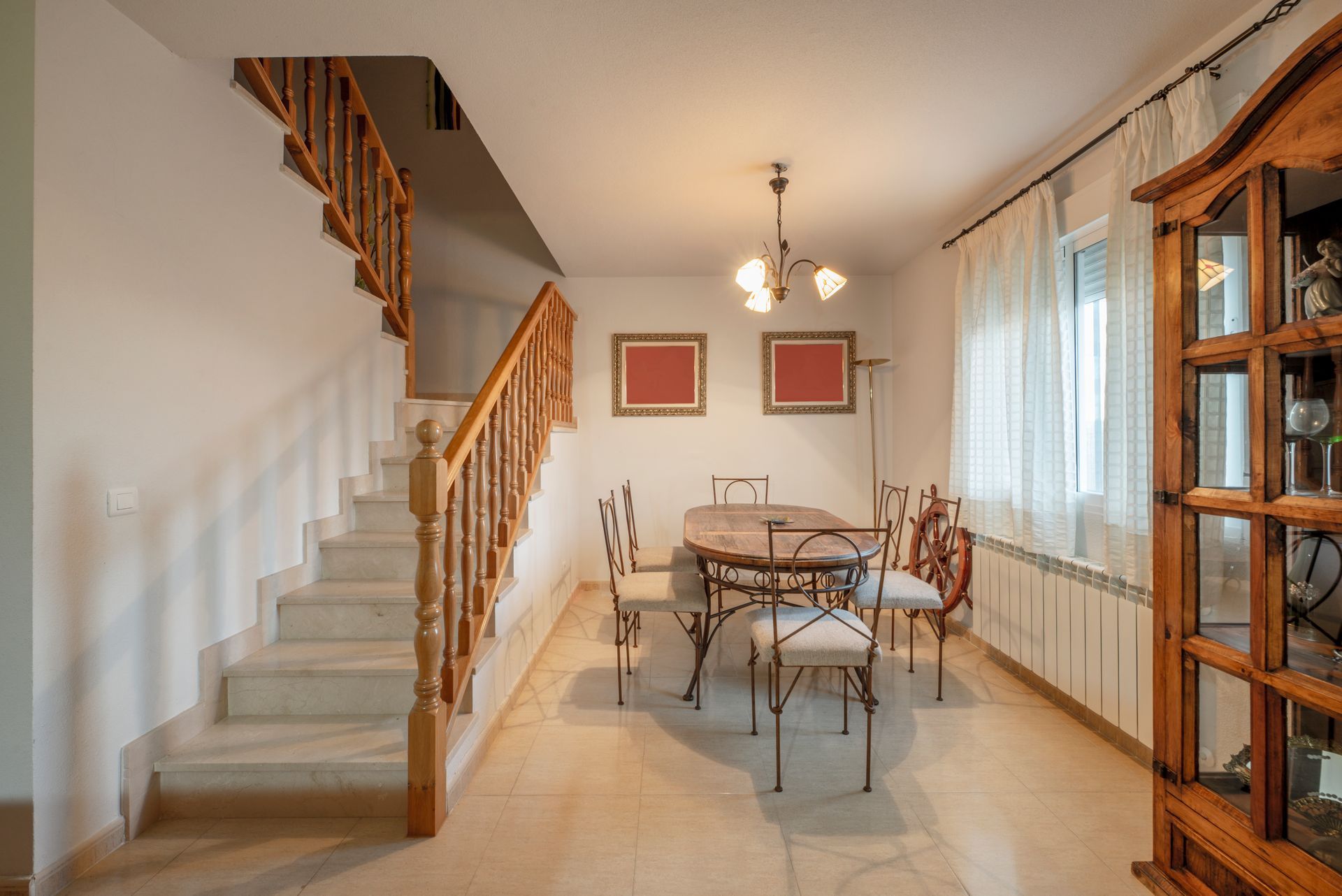 A dining room with a table, chairs, a chandelier, a wooden staircase, and a wood-framed display cabinet.