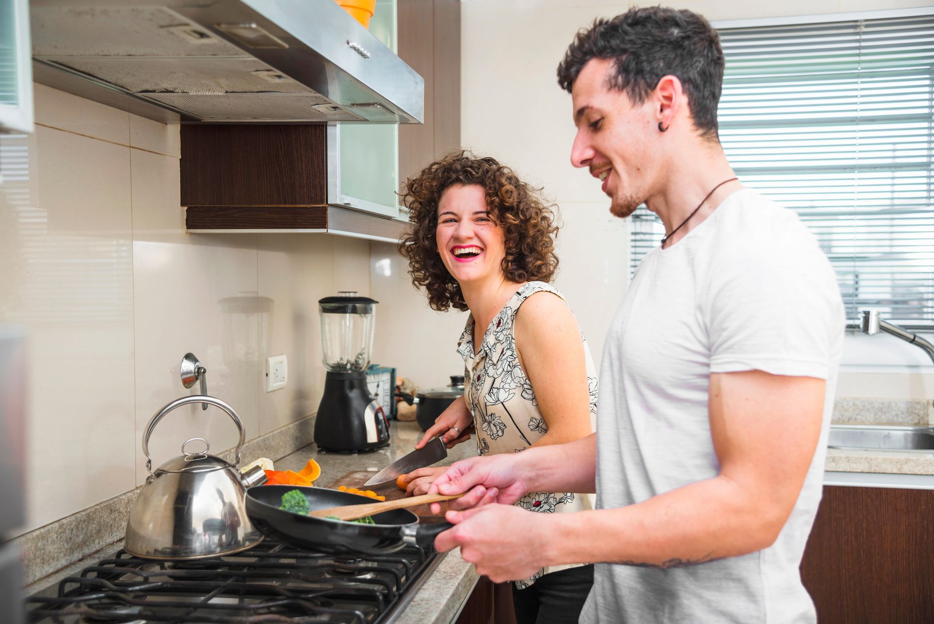 A person with curly hair laughing while cutting vegetables next to another person cooking in a kitchen.