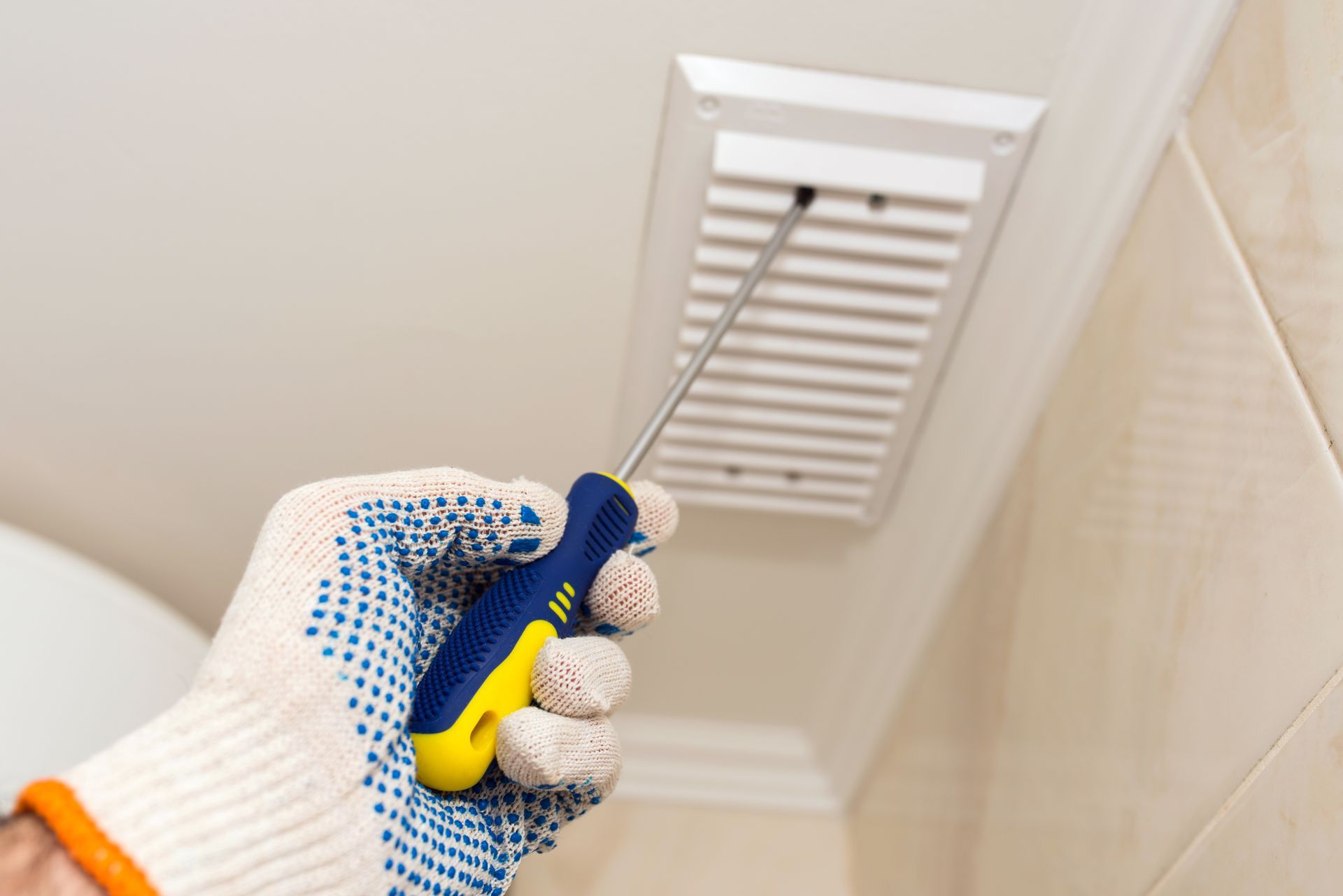Gloved hand using a screwdriver to remove a white vent cover on a ceiling.