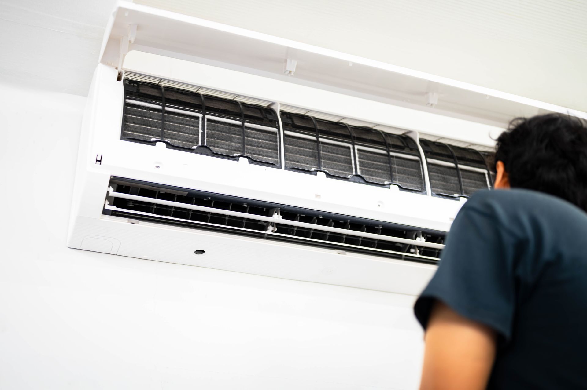 Person looking up at a wall-mounted air conditioner with open vents, set against a white wall.