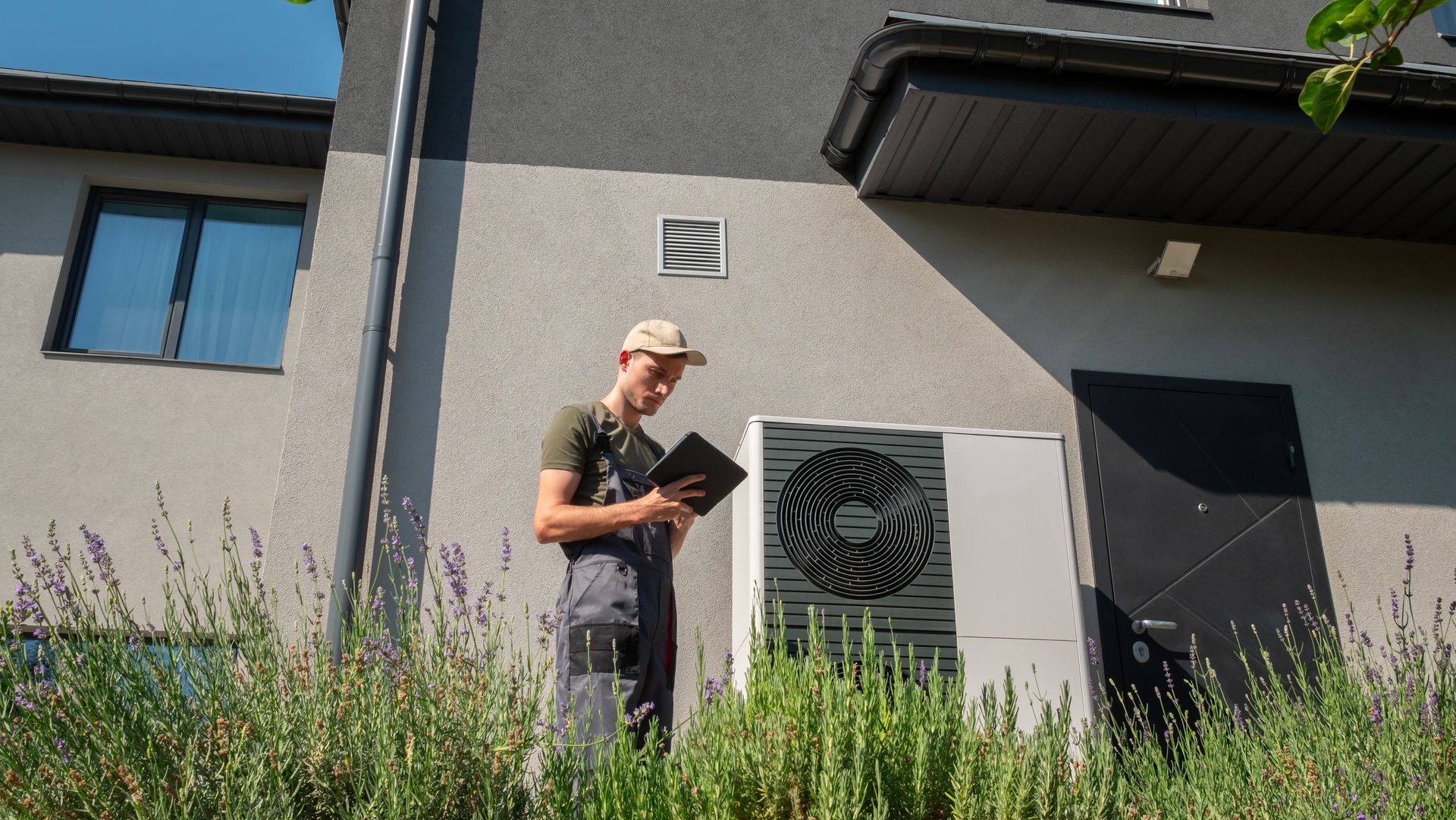 A technician wearing work coveralls and a cap uses a tablet next to an outdoor heat pump unit at a house.