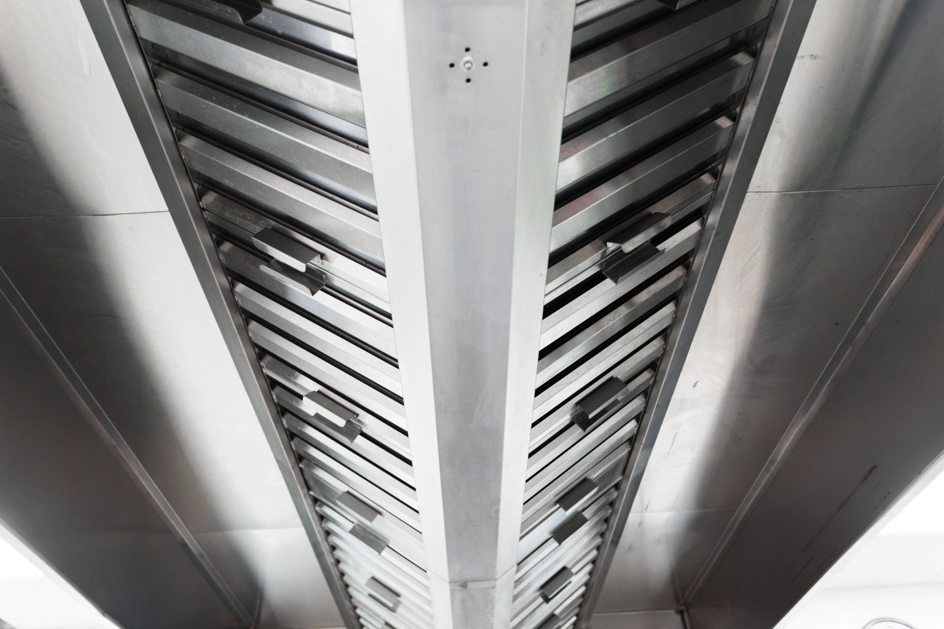 Inside of a stainless steel commercial kitchen hood, with chevron-patterned baffles and a central beam.
