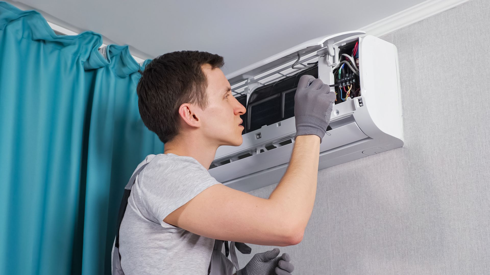 A person in a gray shirt and gloves is working on an air conditioning unit mounted on a wall.