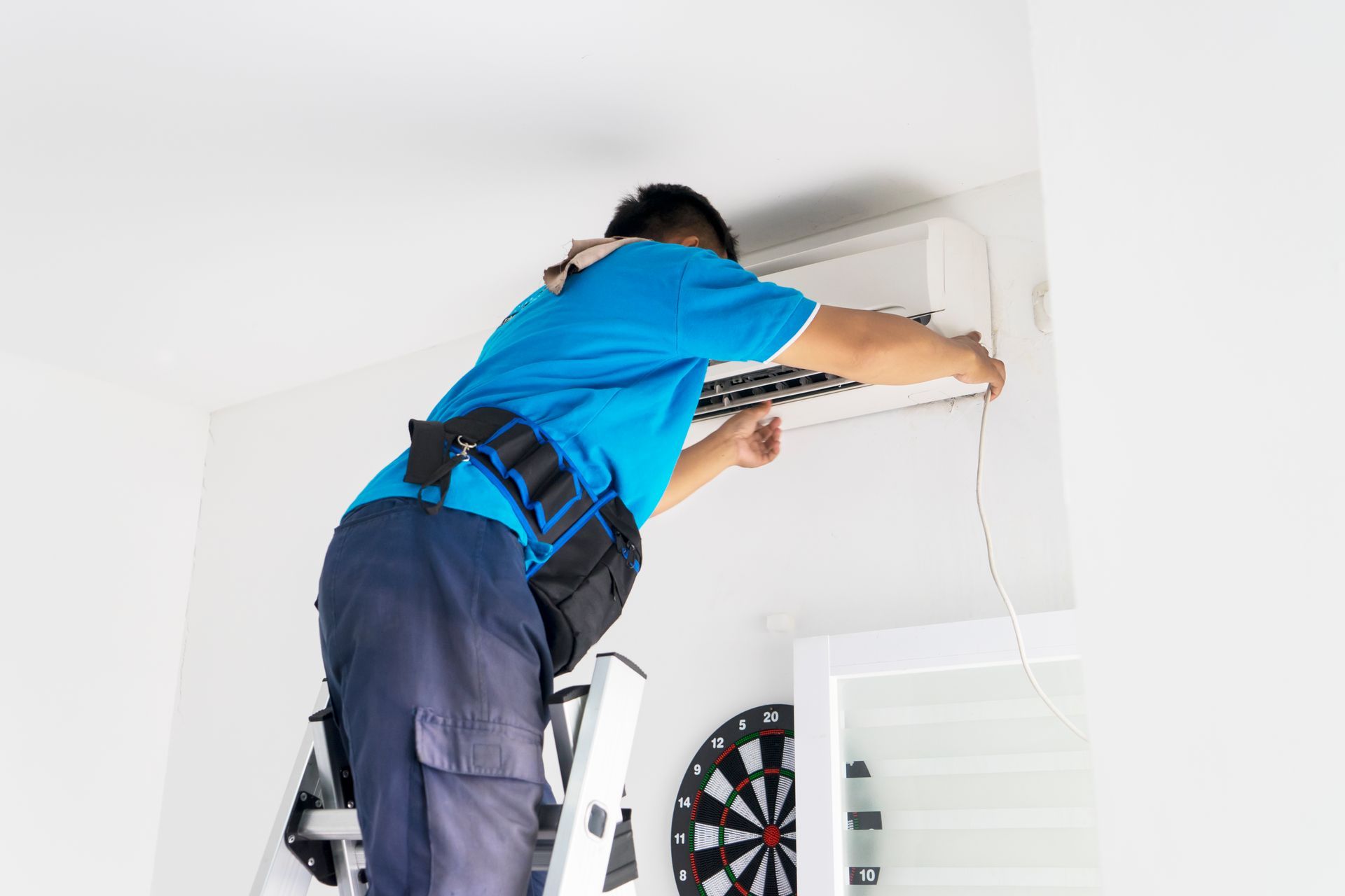 Technician on a ladder, repairing an air conditioning unit on a white wall; dart board visible.