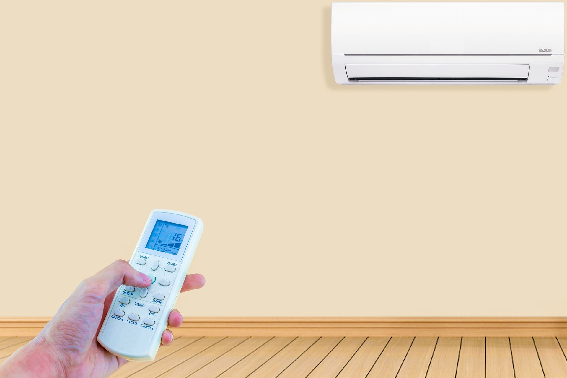 Hand holding AC remote, pointing at wall-mounted air conditioner in a room with wood floor.