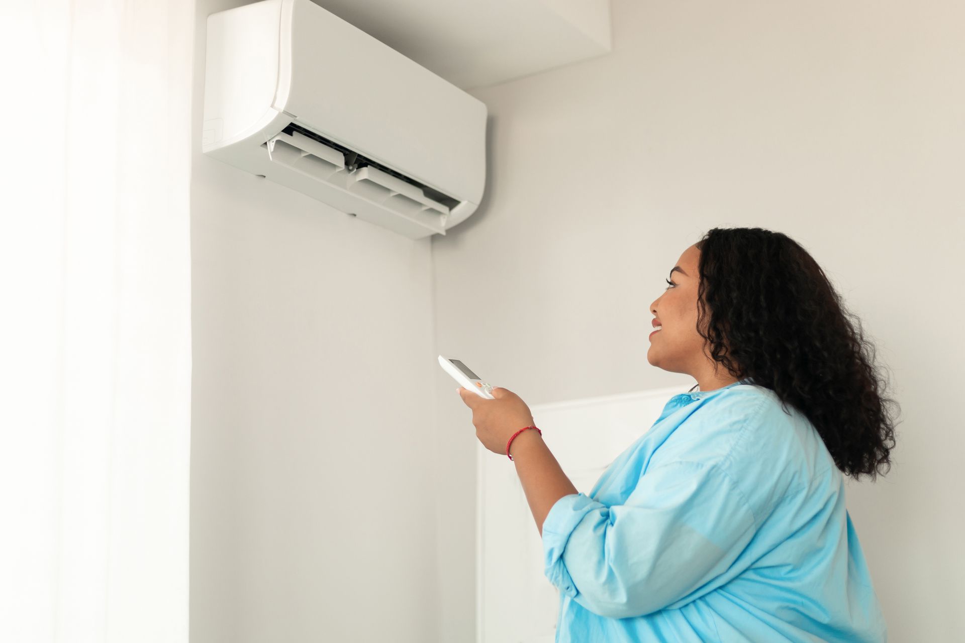 Woman in blue shirt using remote to operate a wall-mounted air conditioner. Bright room, white walls.