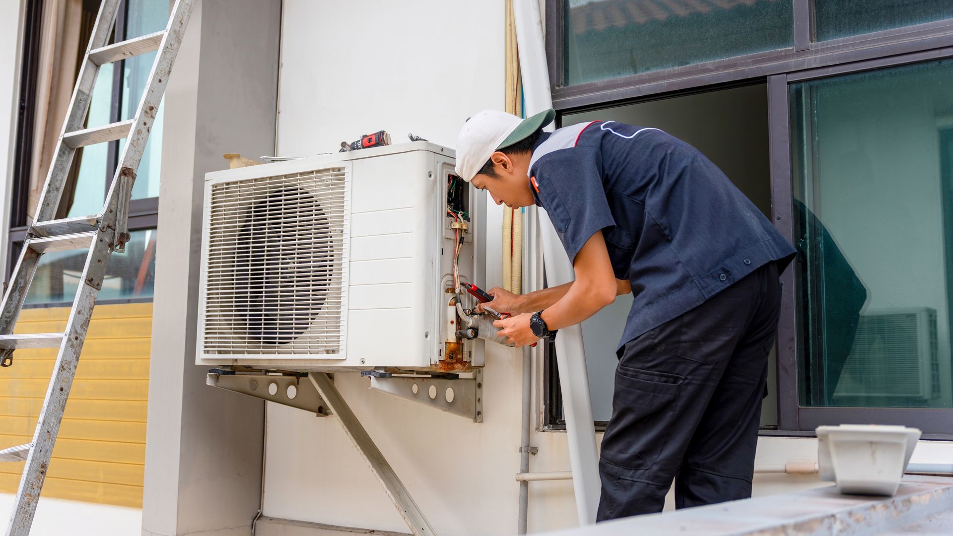 HVAC technician in a white helmet fixing an outdoor air conditioning unit next to a ladder.