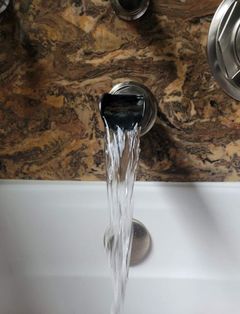 A silver bathtub faucet attached to a brown marble wall, with a steady stream of water flowing into a white tub.