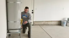 A person wearing a grey hoodie kneels in a garage, inspecting the lower controls of a silver residential water heater.