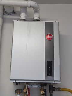 A wall-mounted Rheem tankless water heater with white pipes above and plumbing connections underneath against a white wall.