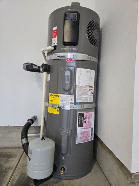 A gray hybrid heat pump water heater with an attached expansion tank, installed in a corner of a garage.