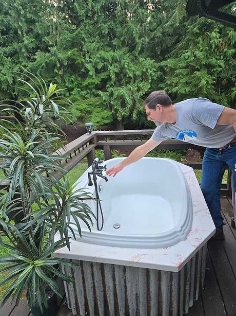 A person in a gray shirt reaches toward a faucet attached to an outdoor bathtub surrounded by a wooden deck and trees.