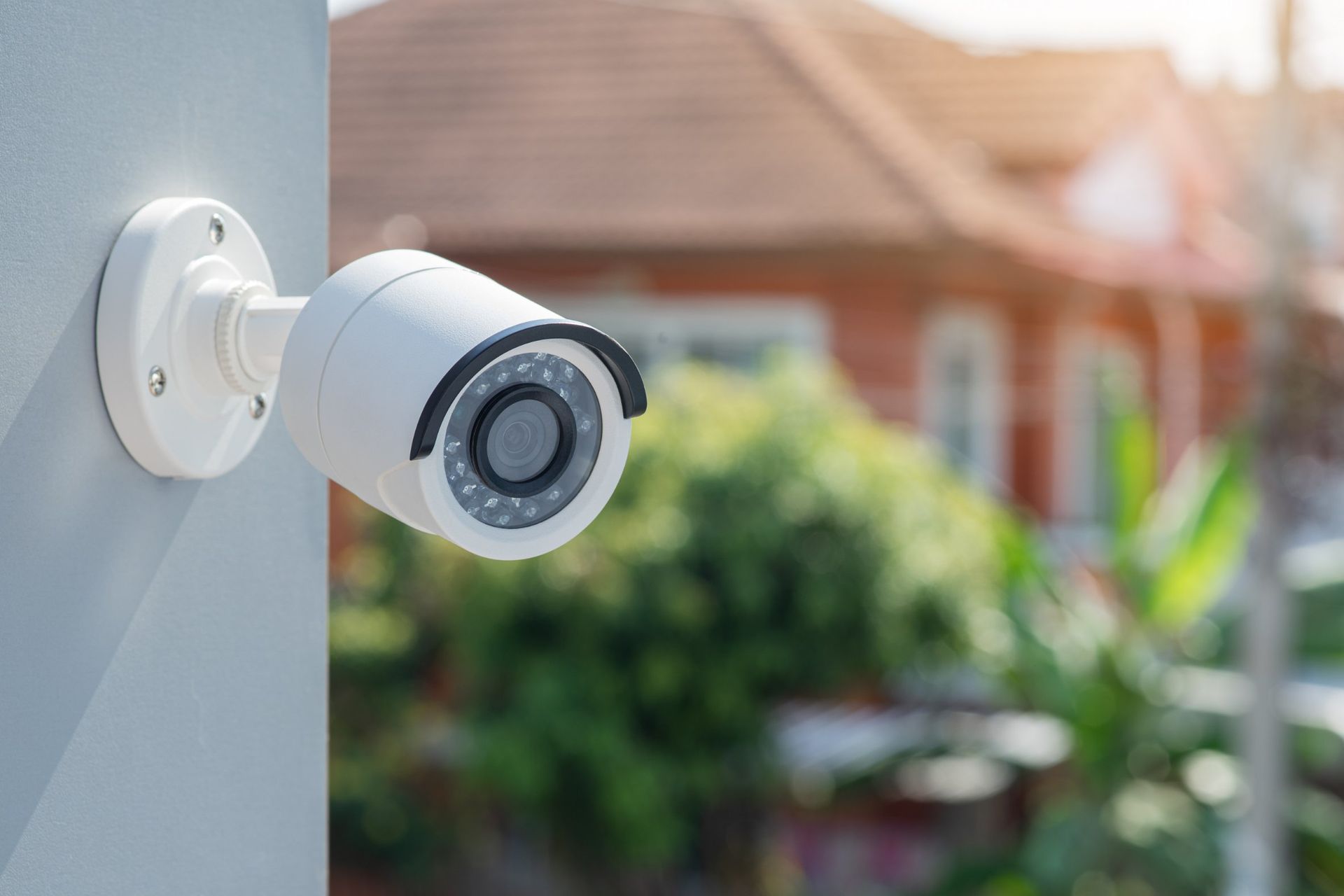 White security camera mounted on a gray wall, overlooking a house with a red brick exterior.