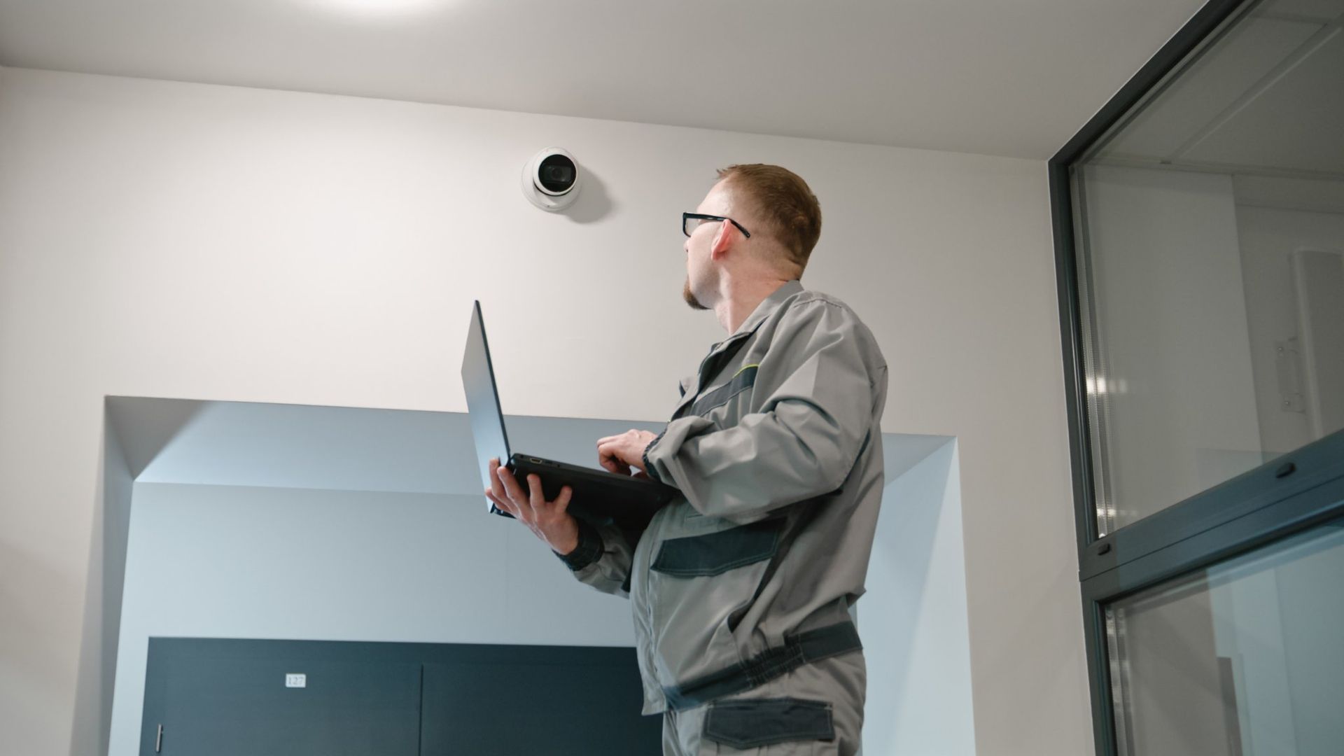 Man in gray work suit with laptop looking up at security camera in hallway.