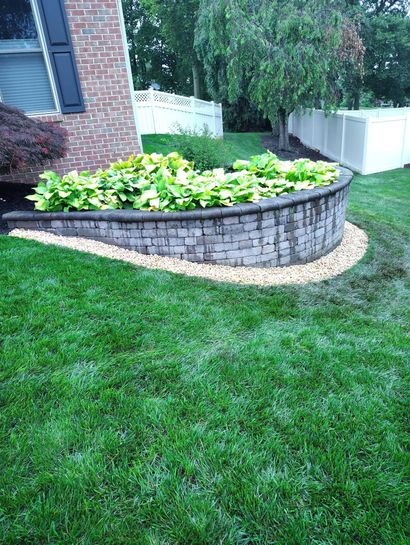 Retaining Walls A curved stone retaining wall filled with lush green plants next to a brick house and green lawn.