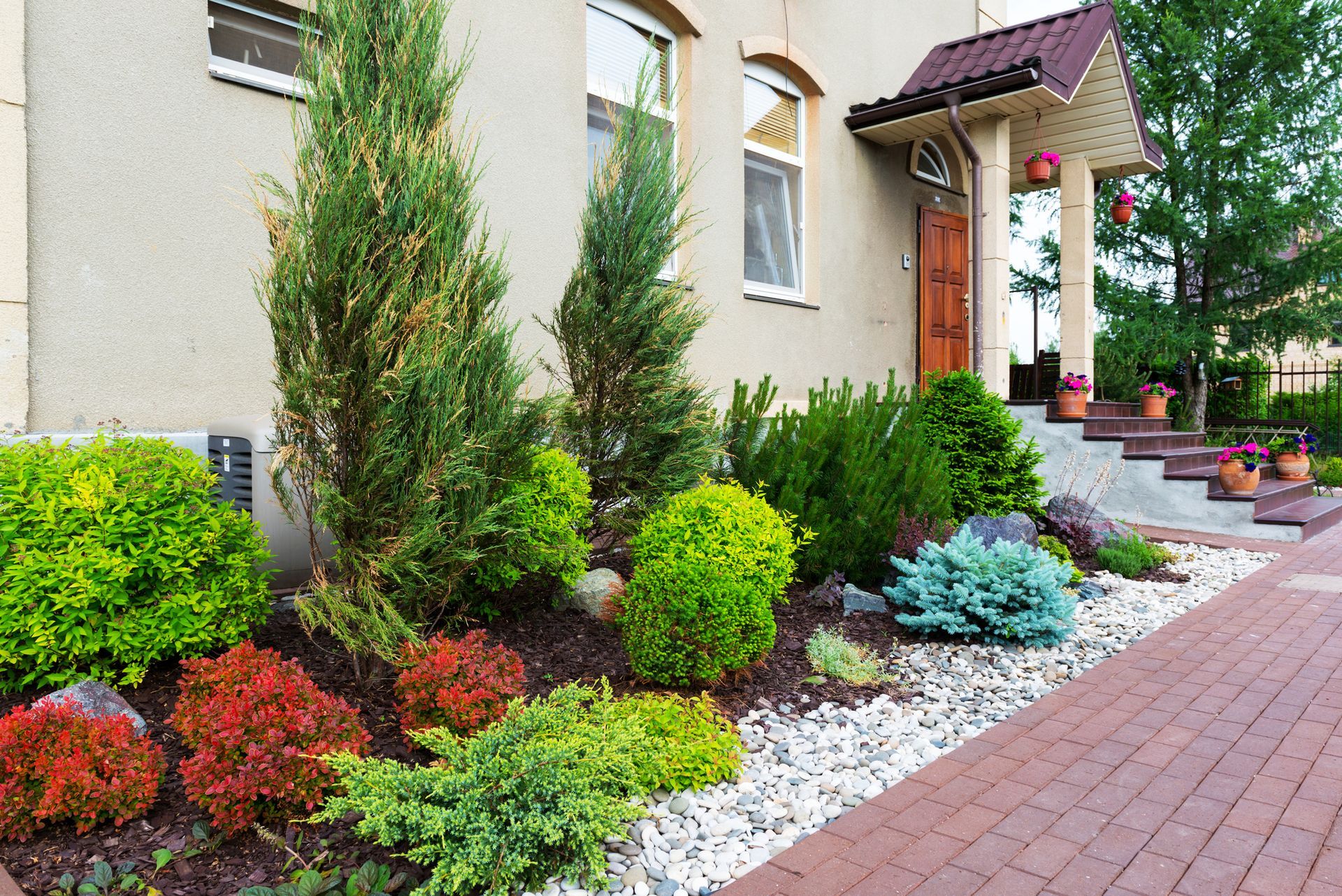 Front yard landscaping with shrubs, white stones, and a brick walkway beside a house entrance
