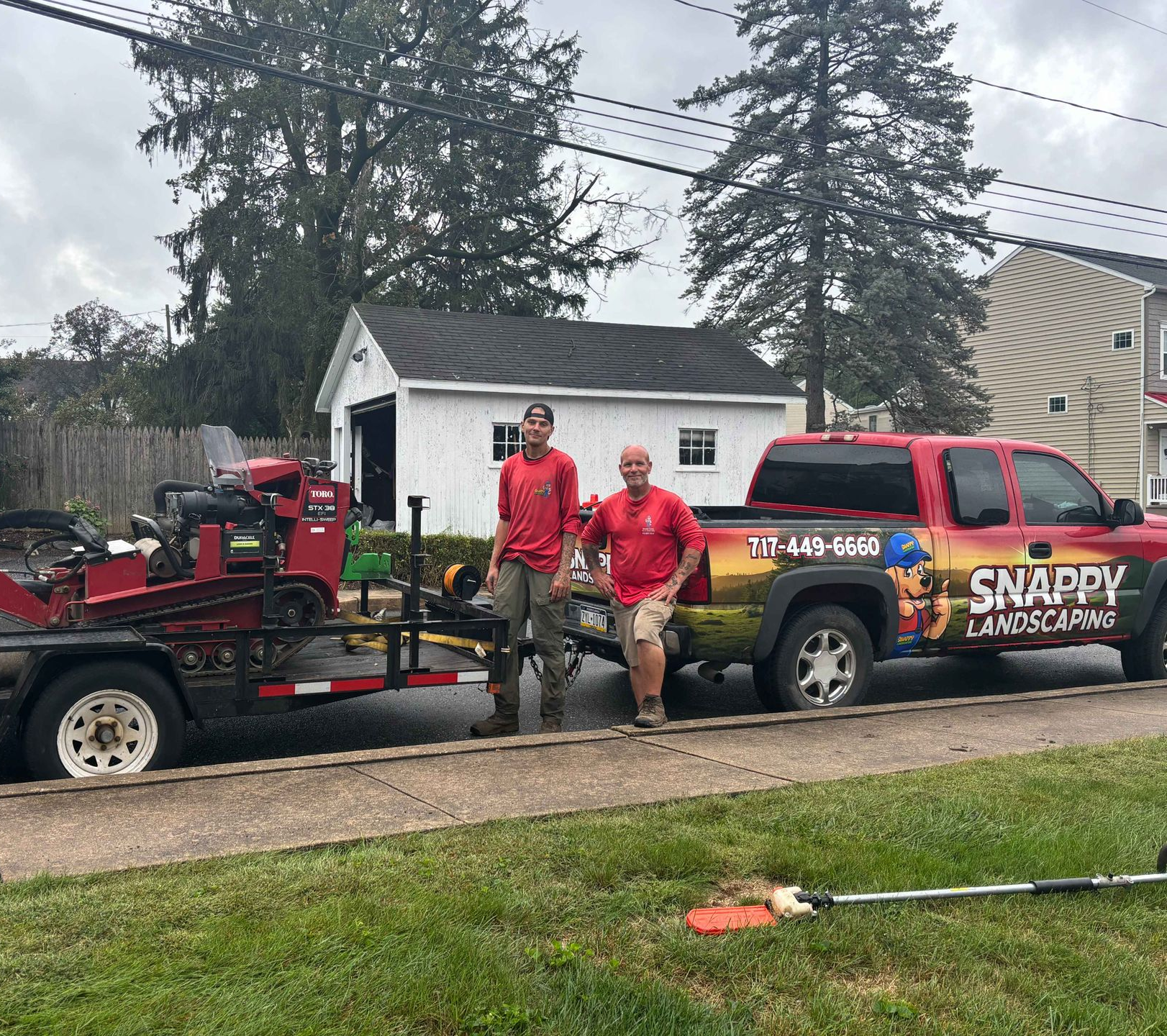 Two men stand by a red truck and trailer with landscaping equipment. 