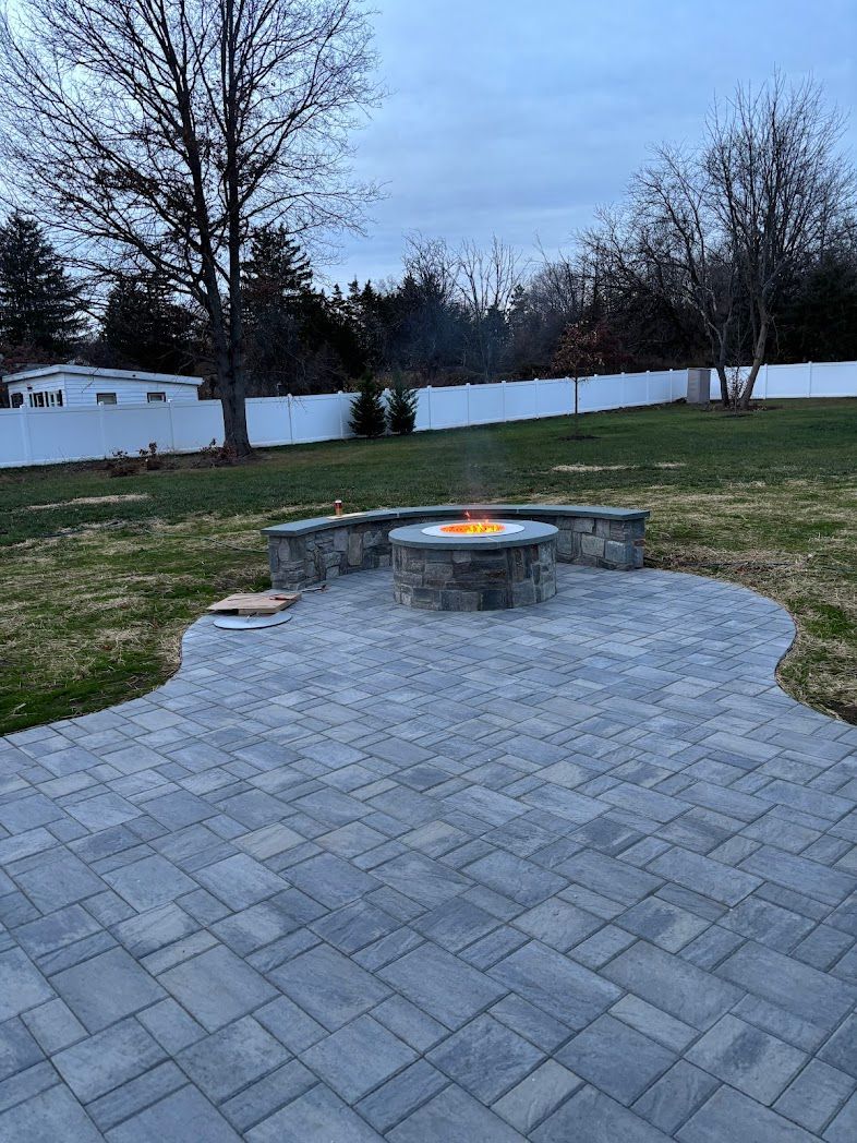 Stone patio with a round fire pit burning at dusk, bordered by grass and a white fence.