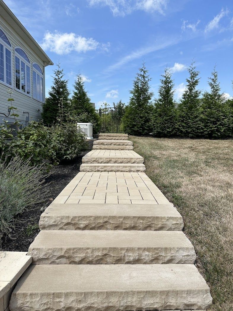 Stone walkway steps through a landscaped yard toward evergreen trees under a blue sky