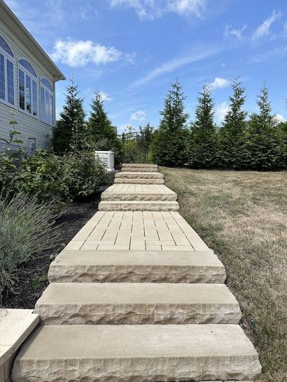 Stone walkway steps through a landscaped yard toward evergreen trees under a blue sky