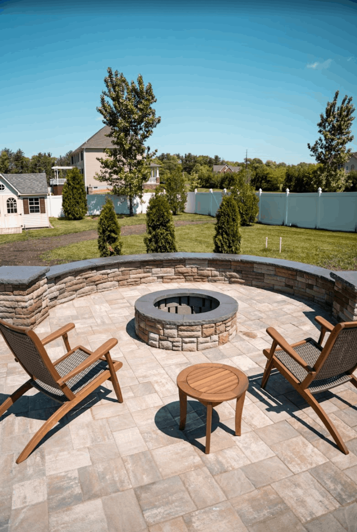 Patio seating around a stone fire pit in a backyard with a lake view and blue sky
